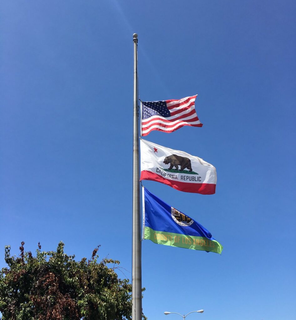 The American flag, State of California Flag, and County of Los Angeles Flag fly at half-staff outside a county building.