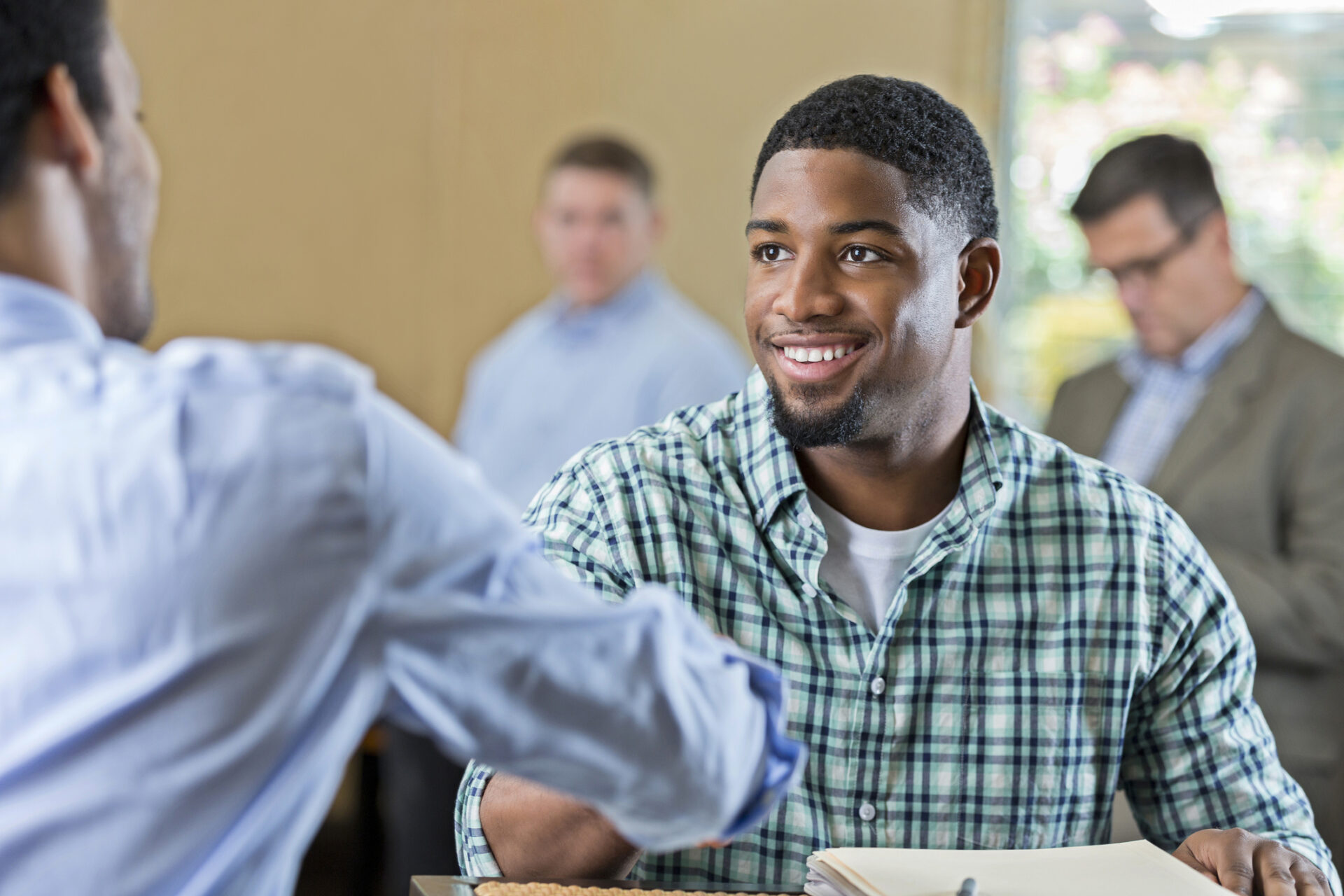 Young African American man smiles while shaking hands with a businessman. The young man is interviewing for a position. He is wearing a plaid shirt and has short black hair and facial hair. People are in the background.